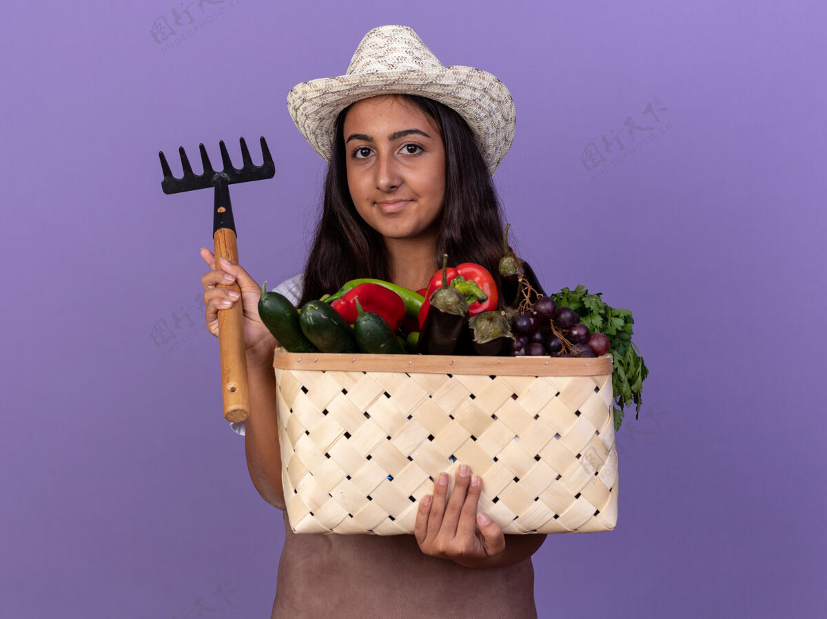 充分穿着围裙 戴着夏帽 拿着装满蔬菜和迷你耙子的小园丁女孩站在紫色的墙上 脸上带着微笑迷你帽子花园