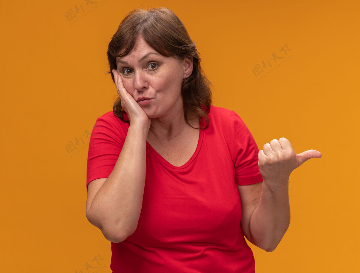 拇指身穿红色t恤的中年妇女惊讶地用拇指指着站在橙色墙上的一边惊人中间指点