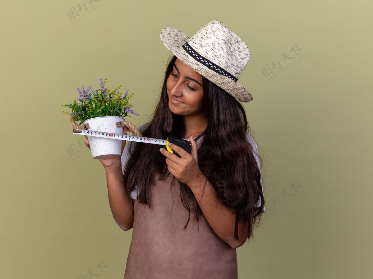 花园穿着围裙 戴着夏帽 拿着卷尺和盆栽植物的年轻园丁女孩站在绿色的墙上 面带微笑地看着植物年轻微笑立场