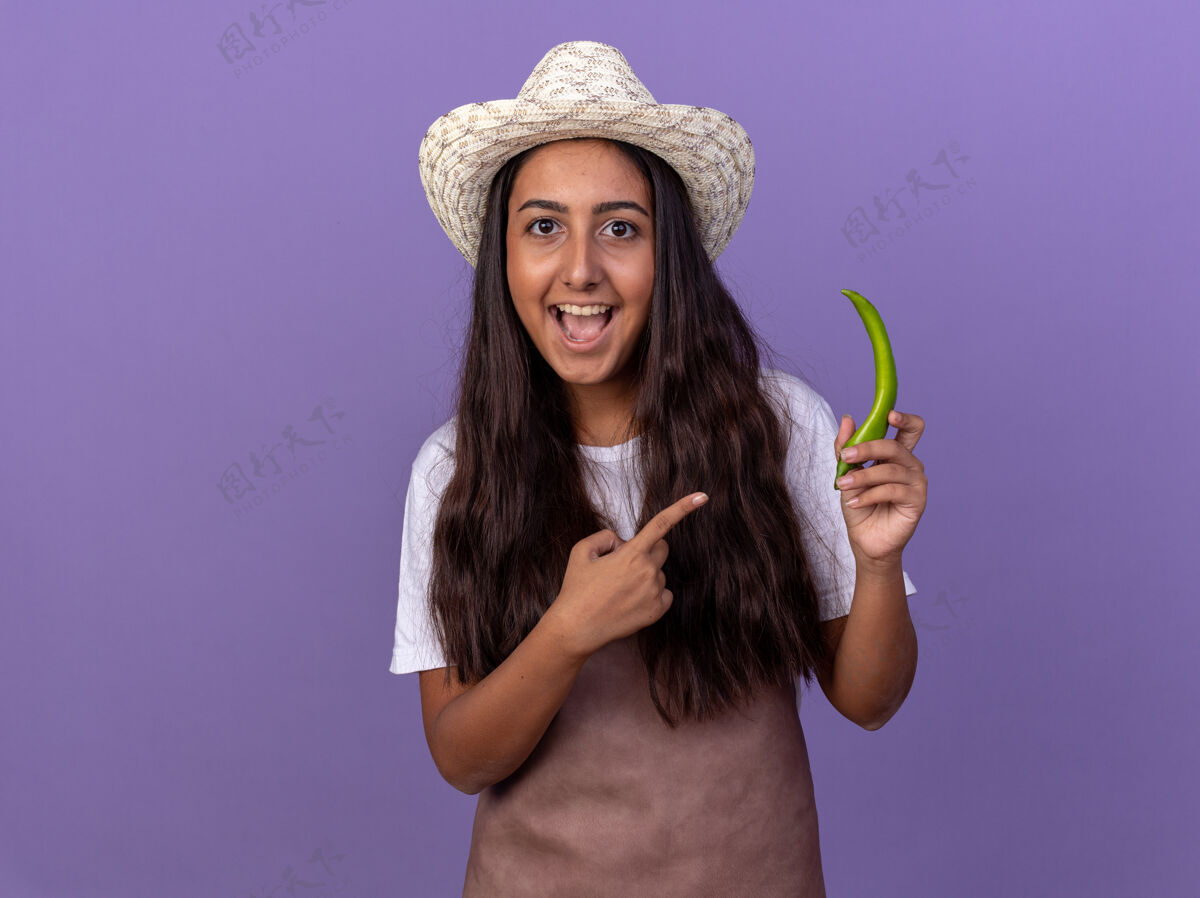 立场穿着围裙 戴着夏帽的年轻园丁姑娘 手拿青椒 食指指着它 站在紫色的墙上 欢快地笑着帽子辣椒辣椒