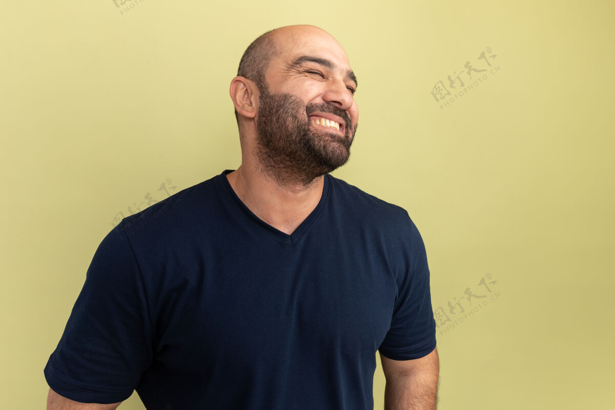 胡须一个留着胡子的男人 穿着黑色t恤 站在绿色的墙上 面带微笑 快乐而积极积极站着一边