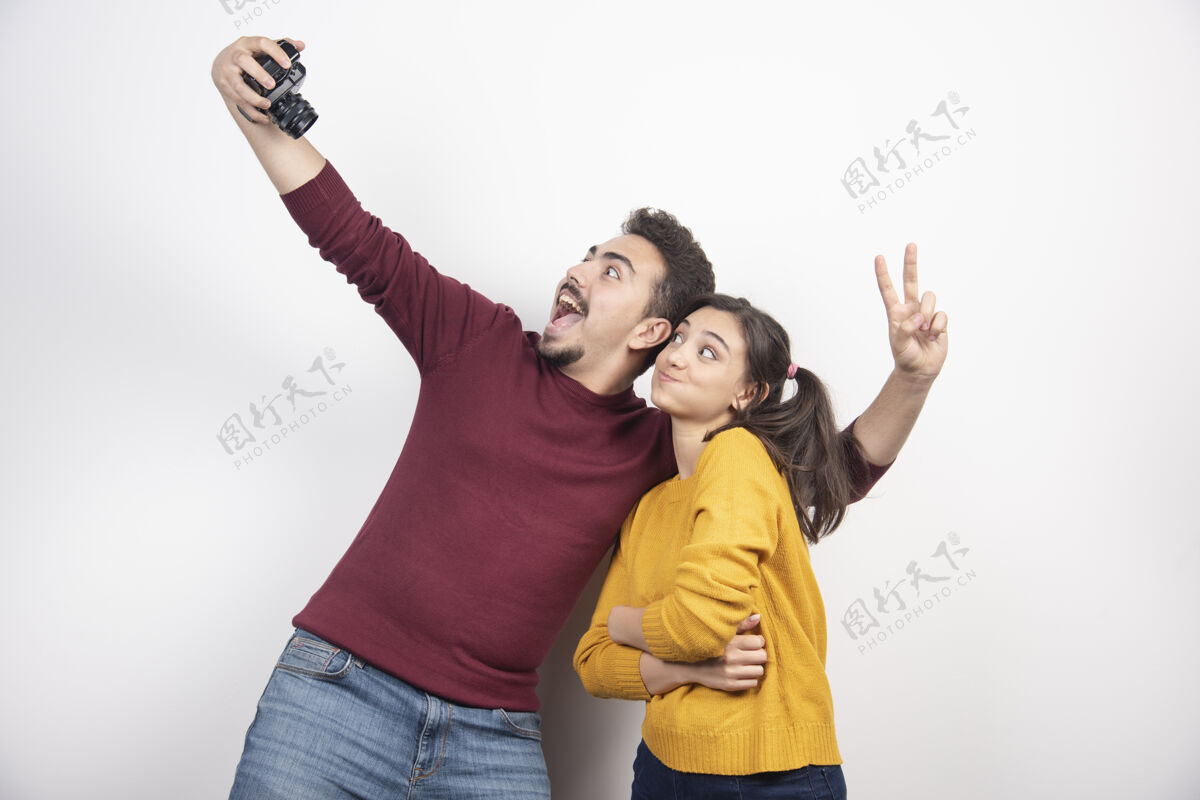 秀一对可爱的情侣拿着相机自拍 在白墙上摆姿势夫妻摄影师微笑