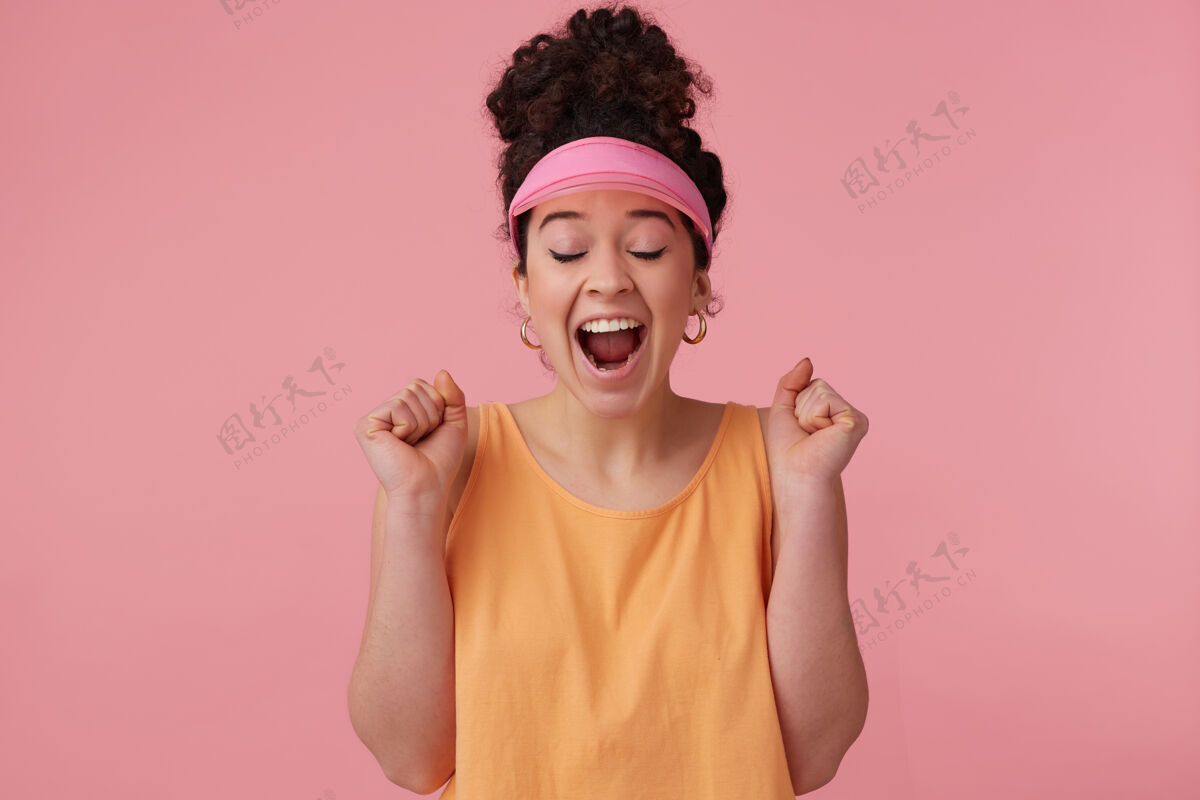 漂亮快乐的 惊讶的女孩 黑色卷发 戴着粉红色的面罩 耳环和橙色的背心 已经化妆 紧握拳头 兴奋地闭上眼睛兴奋心情卷发
