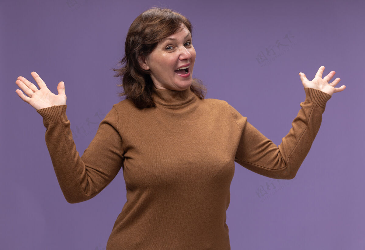 积极穿着棕色高领毛衣的中年妇女快乐而积极地张开双手站在紫色的墙上站立开放女人