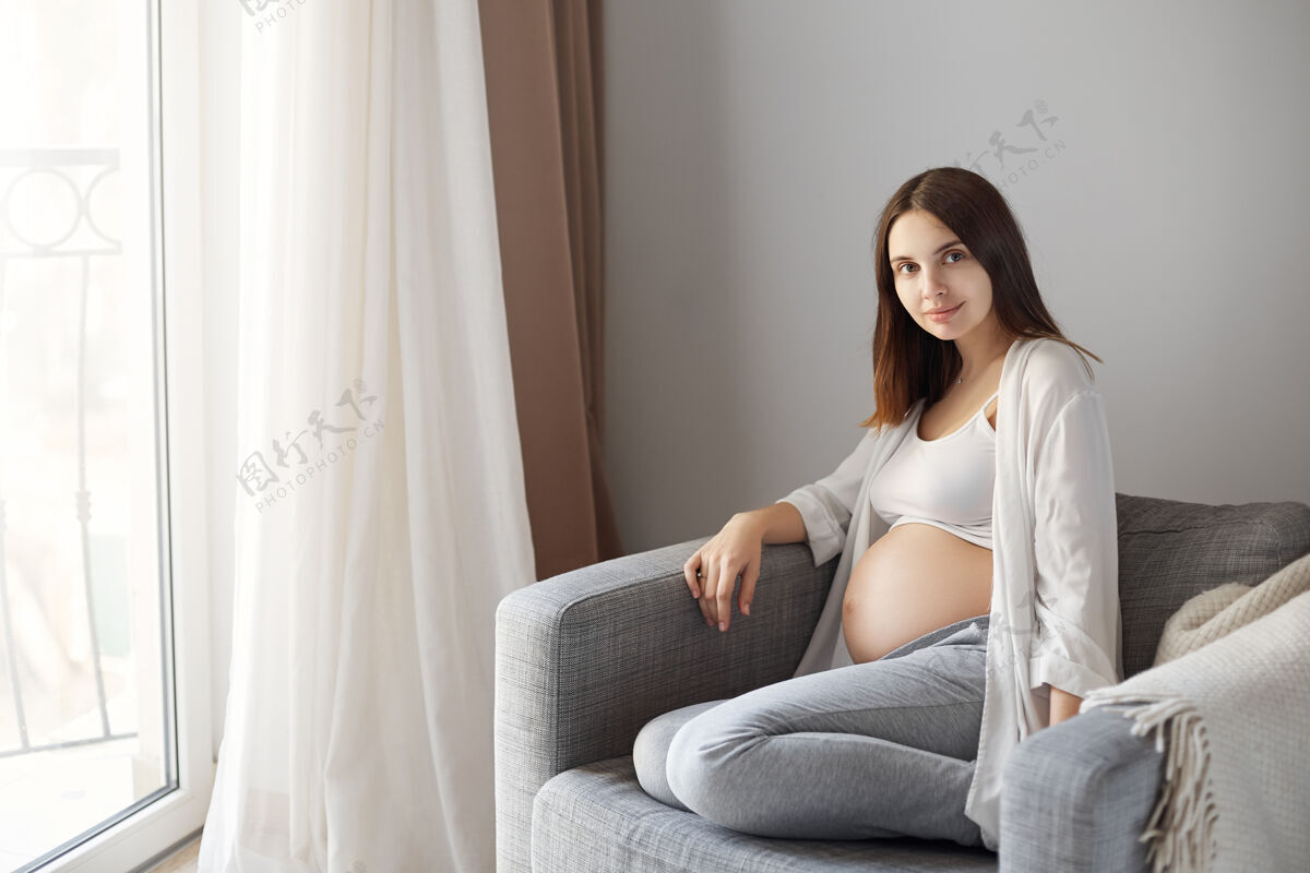 休息丈夫坐在扶手椅上 对着窗边的迷人怀孕妻子拍照 脸上露出可爱温柔的笑容 在等待女儿出生的时候 他平静而快乐室内身体年轻