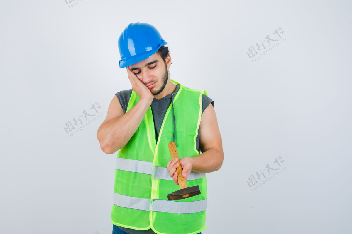 英俊年轻的建筑工人手里拿着锤子 面颊靠在手上 穿着工装 神情沉思 俯视前方沉思而持有