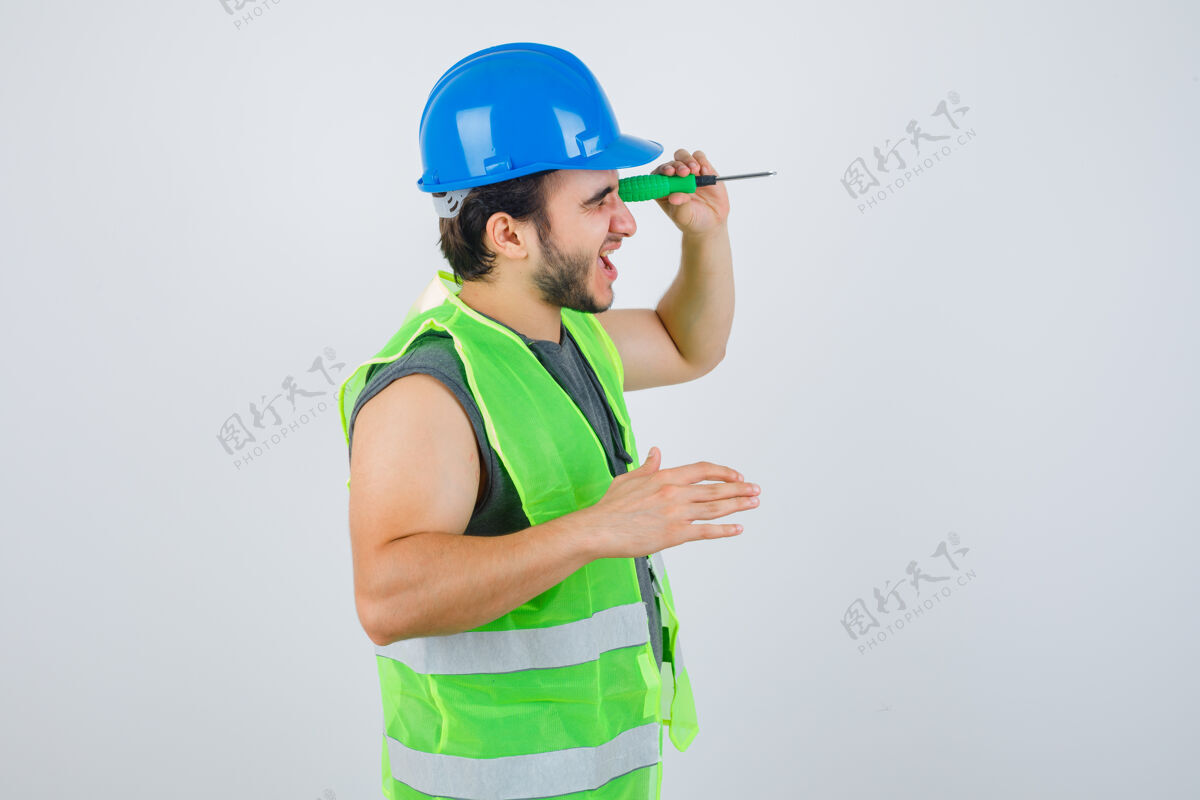 人一幅年轻建筑工人的画像 他穿着制服 假装用螺丝刀偷看一个洞 正看着聚焦的前视图安全螺丝刀孔