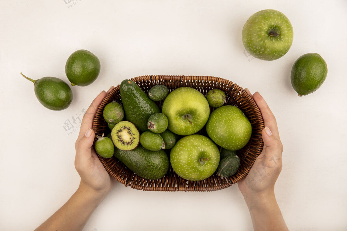 营养女性双手捧着一桶新鲜水果的俯视图 如苹果 菠萝和鳄梨在白色的表面上美味素食新鲜