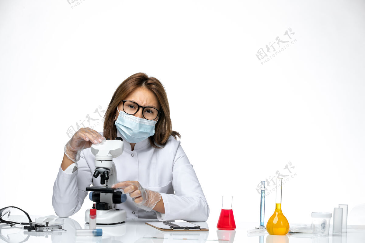 药品前视图女性医生穿着白色的医疗服和面具 由于科维德使用显微镜在光白色空间医疗显微镜套装