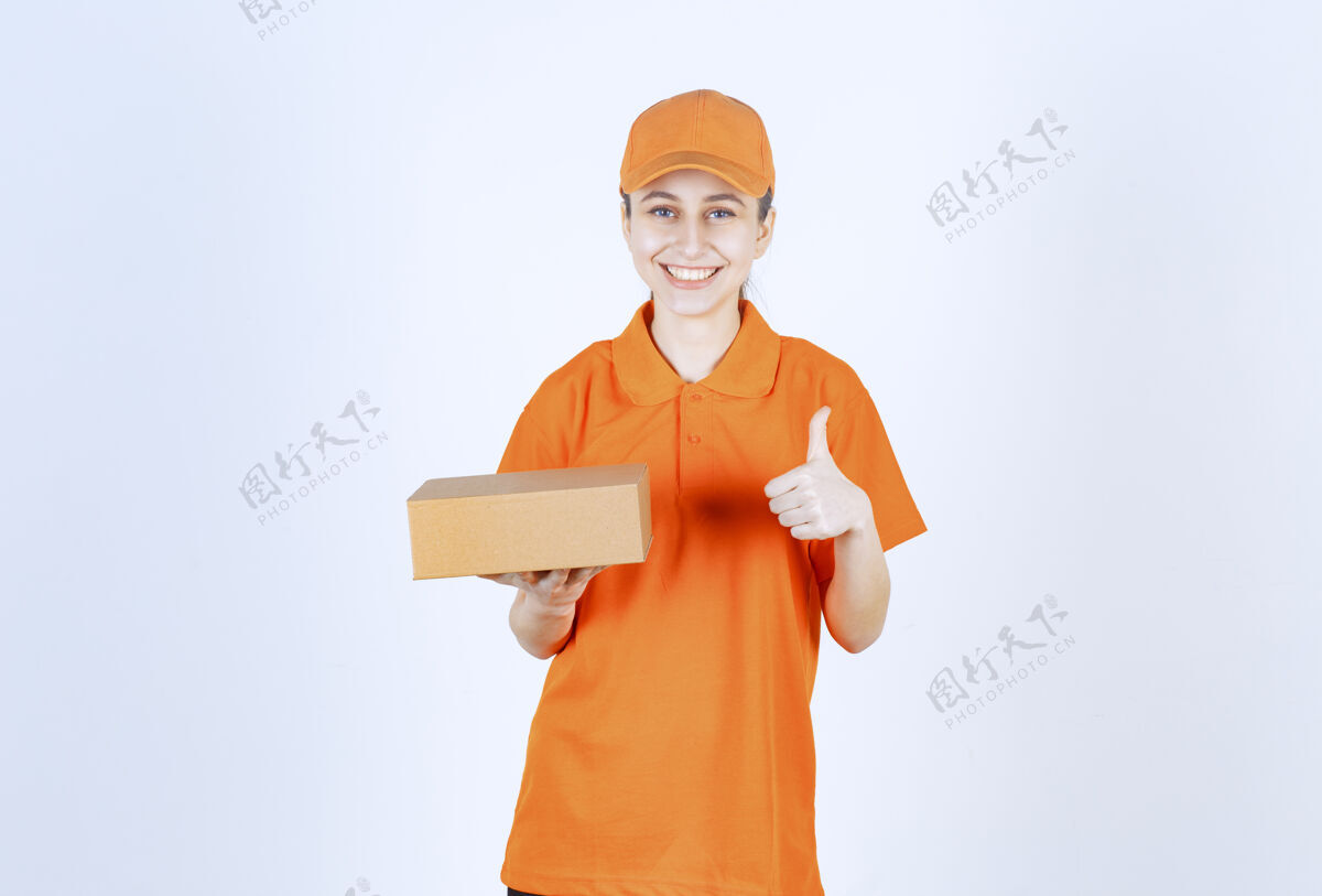 成人穿着黄色制服的女快递员拿着一个纸板箱 手上有肯定的手势强壮订单工人