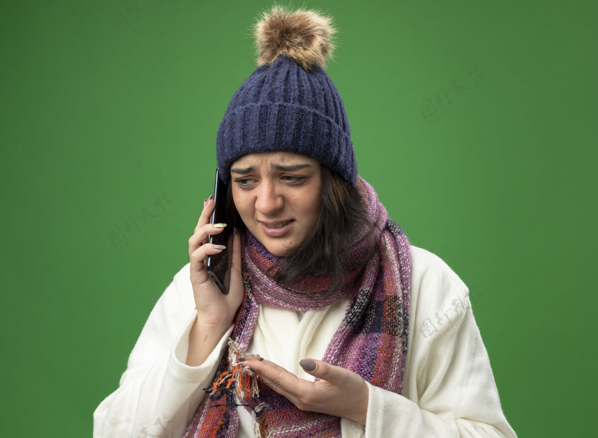 空穿着长袍 戴着冬天的帽子 戴着围巾的年轻白种人生病的女孩在打电话 空手看着隔离在绿墙上的一边电话不愉快穿
