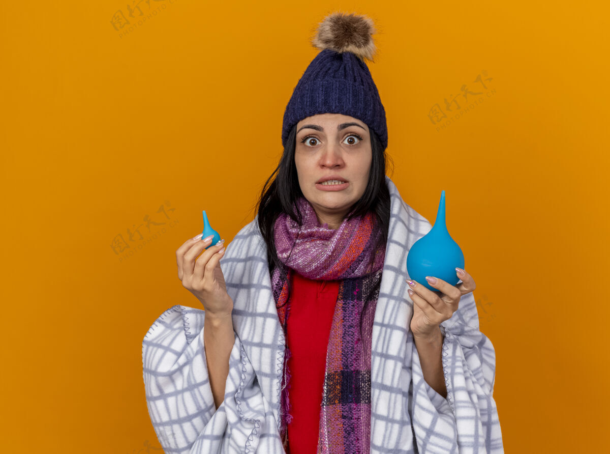 疾病印象深刻的年轻白种人生病的女孩戴着冬天的帽子和围巾包裹在格子布举行灌肠隔离在橙色的墙壁与复制空间格子围巾穿