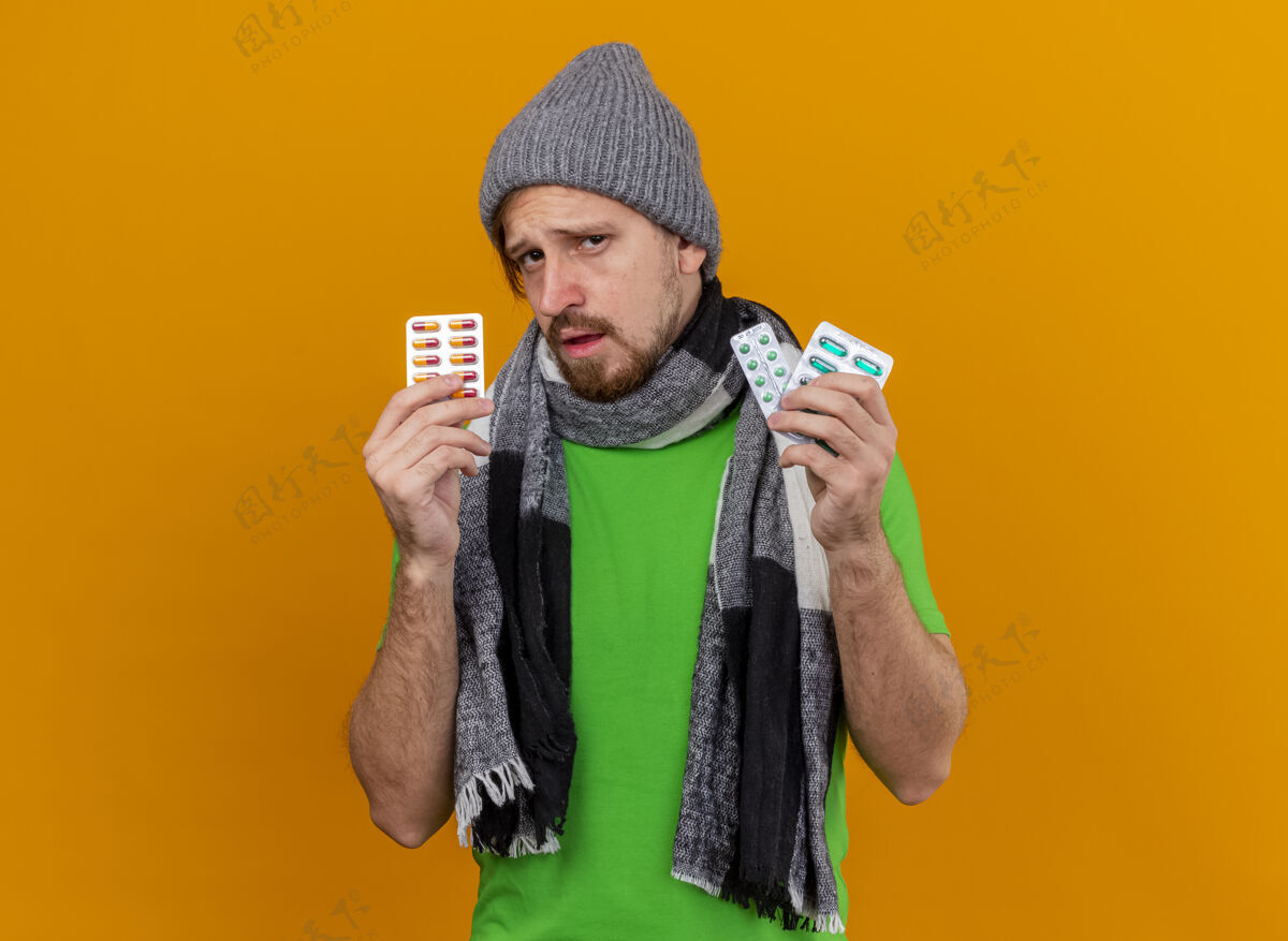 包自信的年轻英俊的斯拉夫病夫戴着冬天的帽子和围巾 手里拿着几包胶囊 隔离在橙色的墙上 留着复印空间斯拉夫围巾年轻