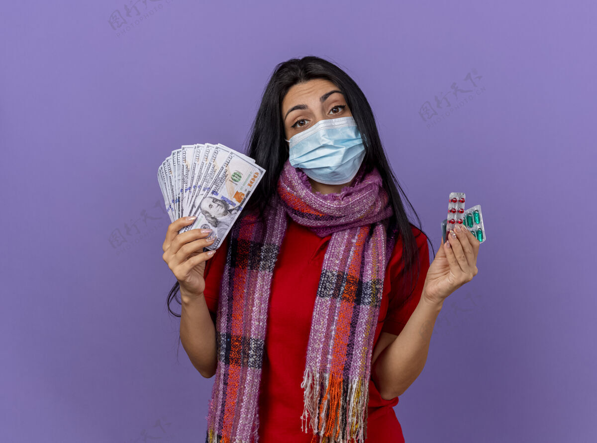 钱自信的年轻白种女孩戴着面具 戴着围巾 手里拿着钱和几包胶囊 隔离在紫色的墙上 留着复印空间围巾胶囊年轻