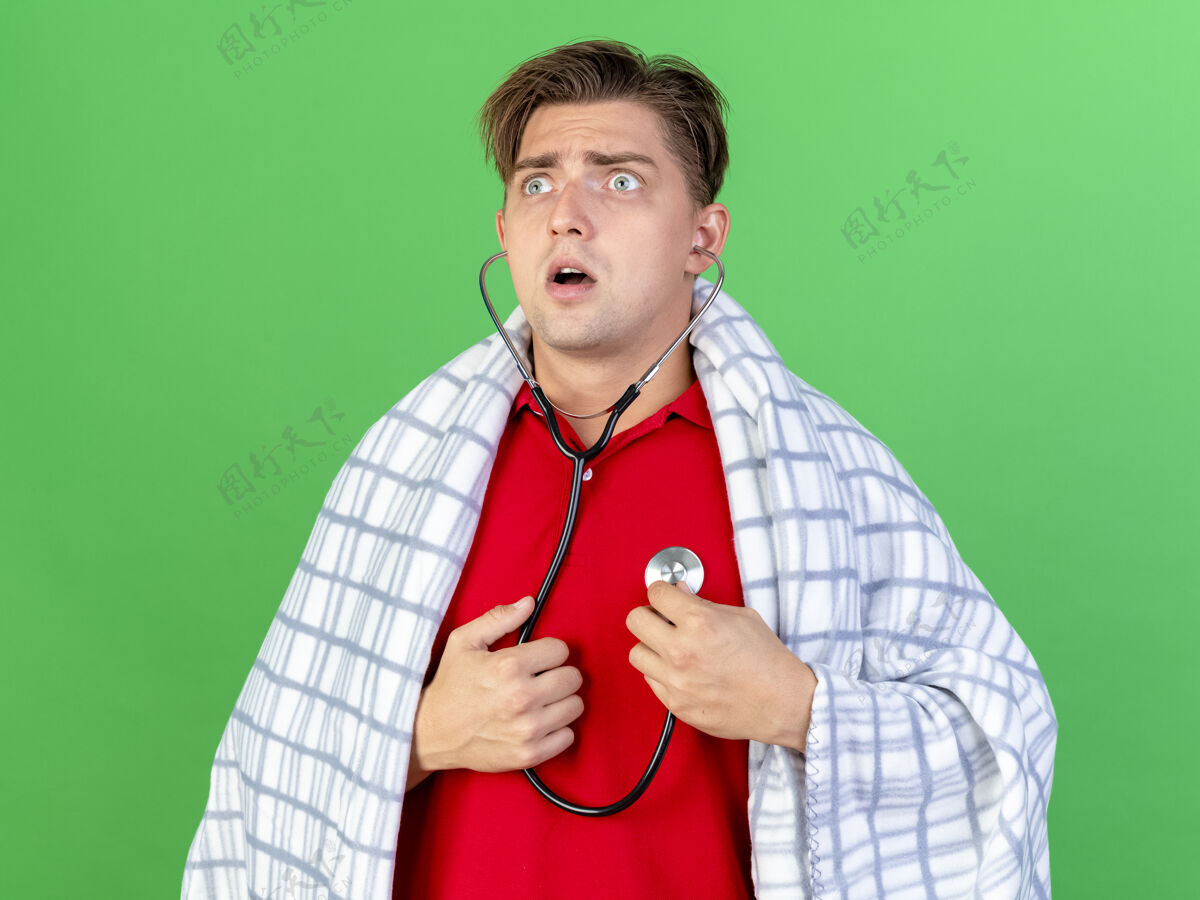 侧身年轻帅气的金发病患戴着听诊器听着自己的心跳看着隔离在绿墙上的一面男人年轻听诊器