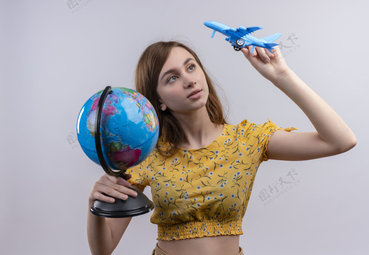 抱着年轻女孩拿着地球仪和模型飞机 看着孤立的白色墙上的模型飞机飞机年轻人女人