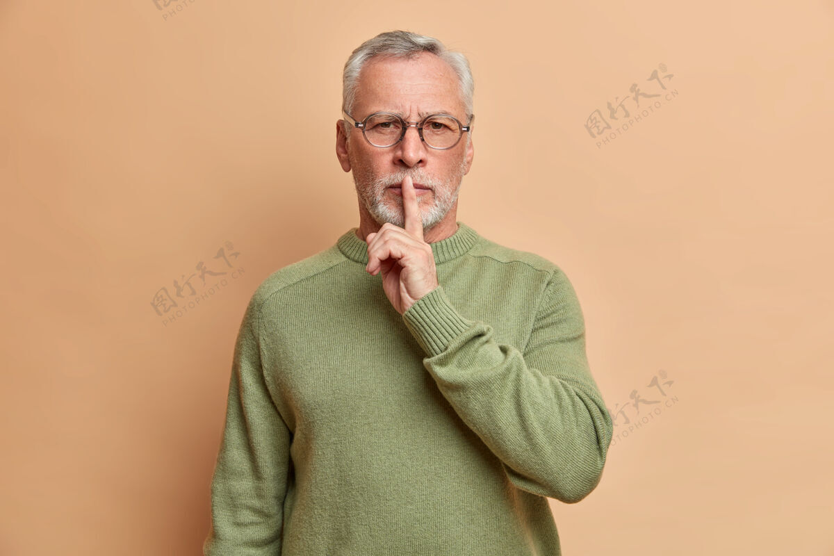 成熟胡子灰白头发的男人严肃地看着前面做了个安静的手势要求保持沉默自信地看着前面戴着透明眼镜和休闲套头衫隔着棕色的墙男人眼镜套头衫