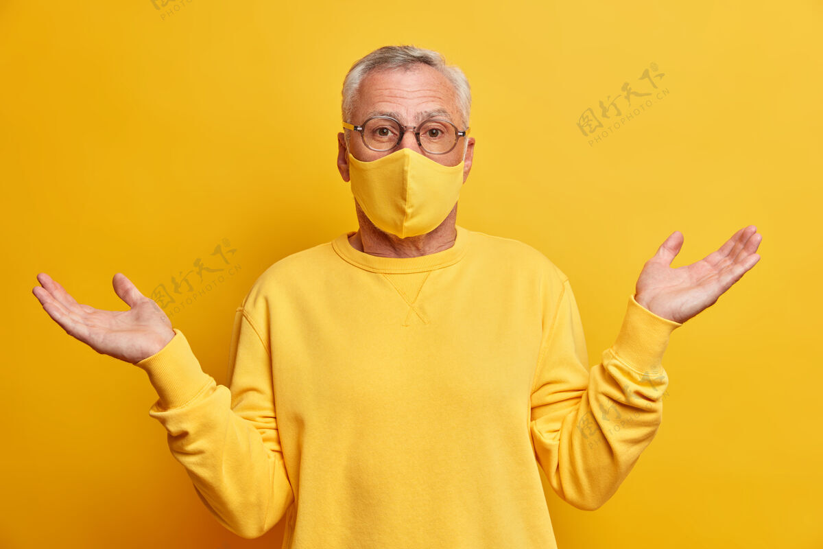 老年人不知情的白发男子不知道发生了什么事传播手掌和站在黄色生动的墙壁困惑戴着防护面具在冠状病毒大流行跳伞个人退休