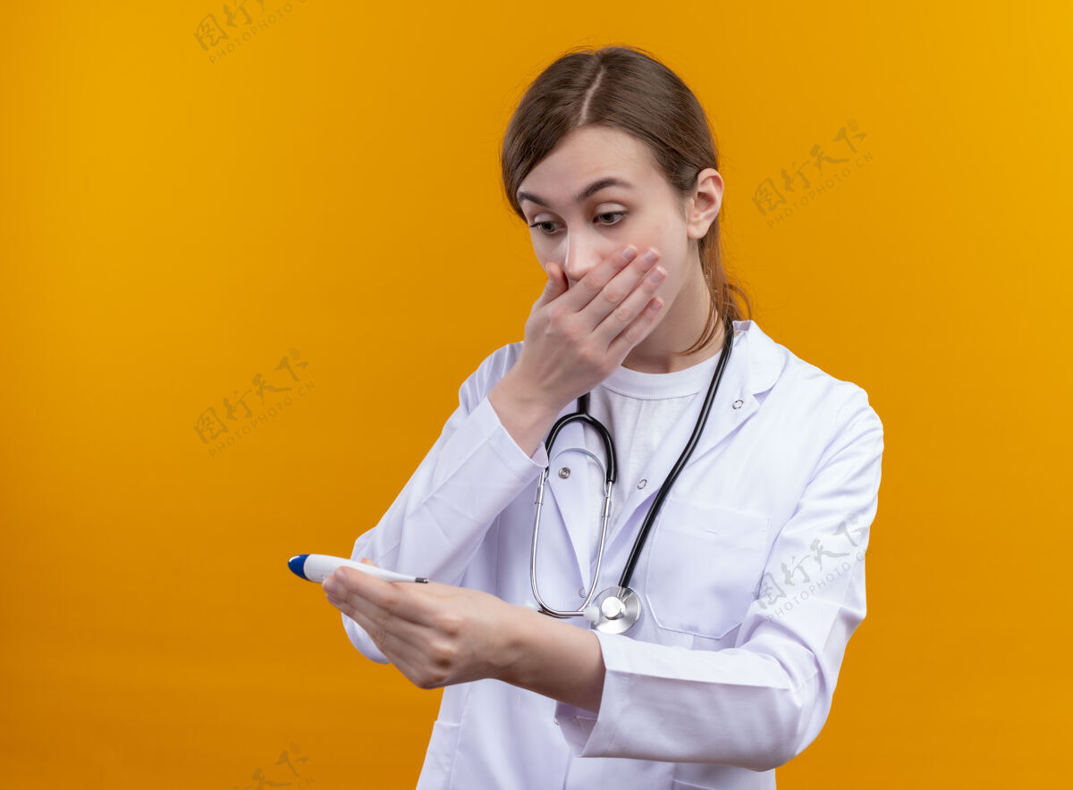 女人惊讶的年轻女医生穿着医用长袍 手持听诊器 手放在口上拿着温度计 在隔离的橙色墙壁上看着它 还有复印空间手体温计年轻人