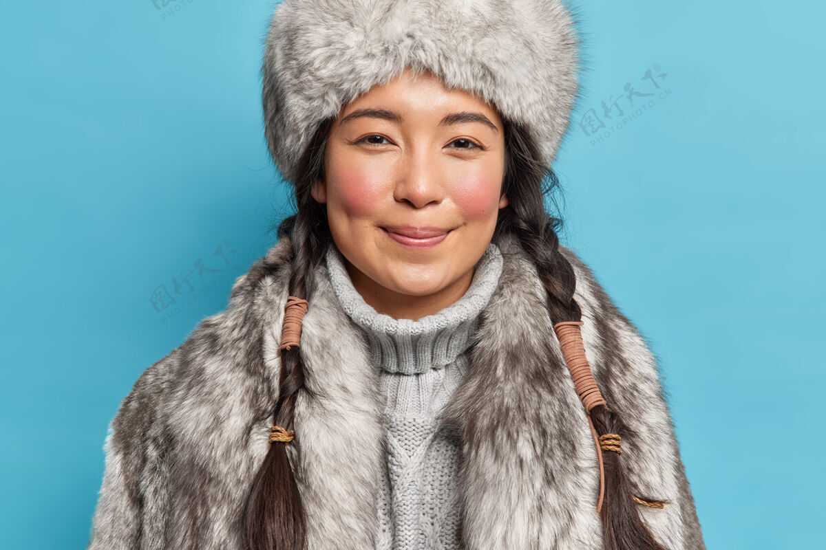 姿势满意的西伯利亚年轻女子 留着两条辫子 红润的脸颊 在寒冷的极地天气里 在蓝色的墙壁上 对着连衣裙露出愉快的微笑毛衣时尚欢呼