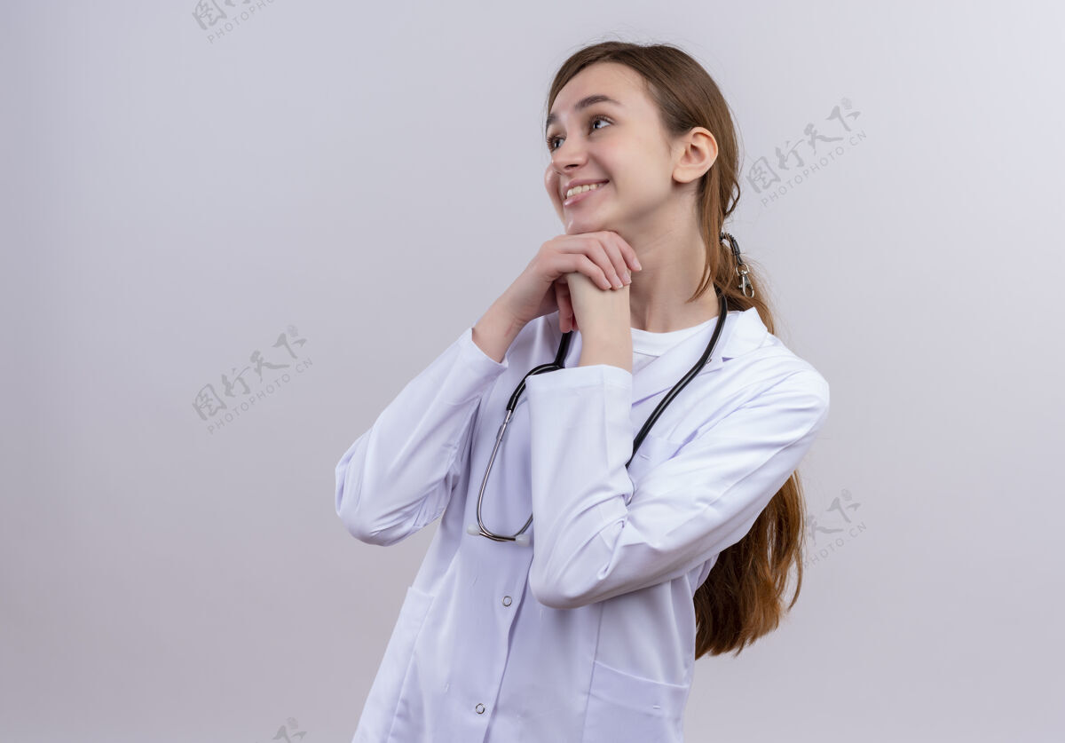 年轻面带微笑的年轻女医生 穿着医用长袍 手持听诊器 将手放在下巴下面 放在隔离的白色墙壁上 留有复印空间听诊器穿长袍