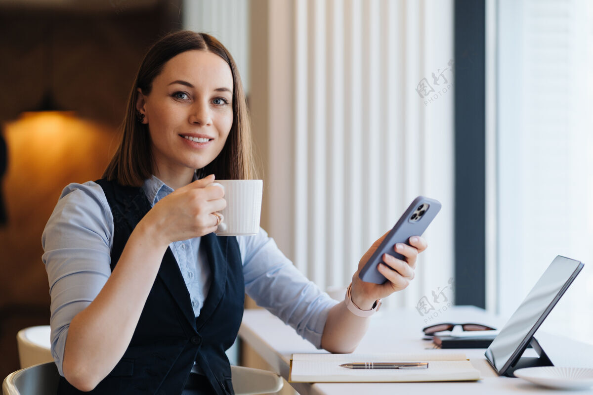 工作年轻迷人的女人一边用智能手机发短信 一边独自坐在咖啡馆喝咖啡 一边用手机聊天坐着通讯使用