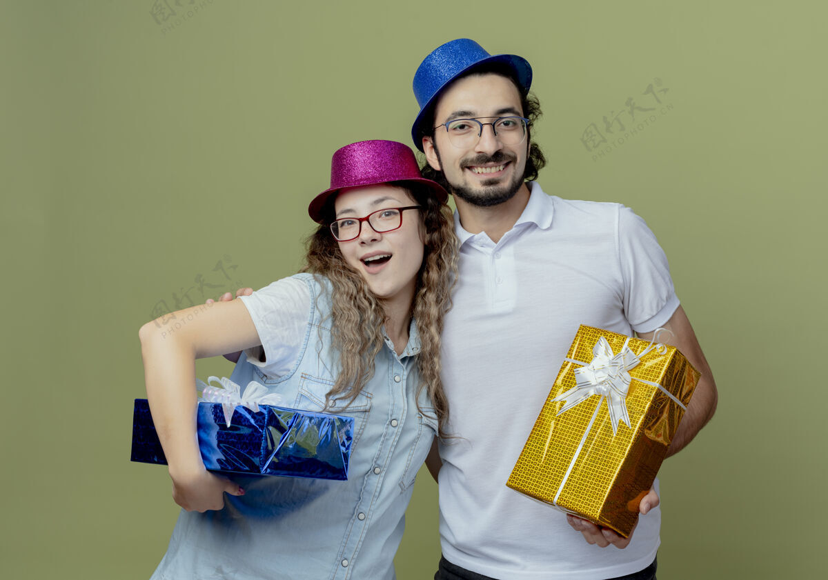 年轻微笑着的年轻夫妇戴着粉色和蓝色的帽子拥抱在一起 手拿着橄榄绿墙上孤立的礼盒帽子每个人盒子