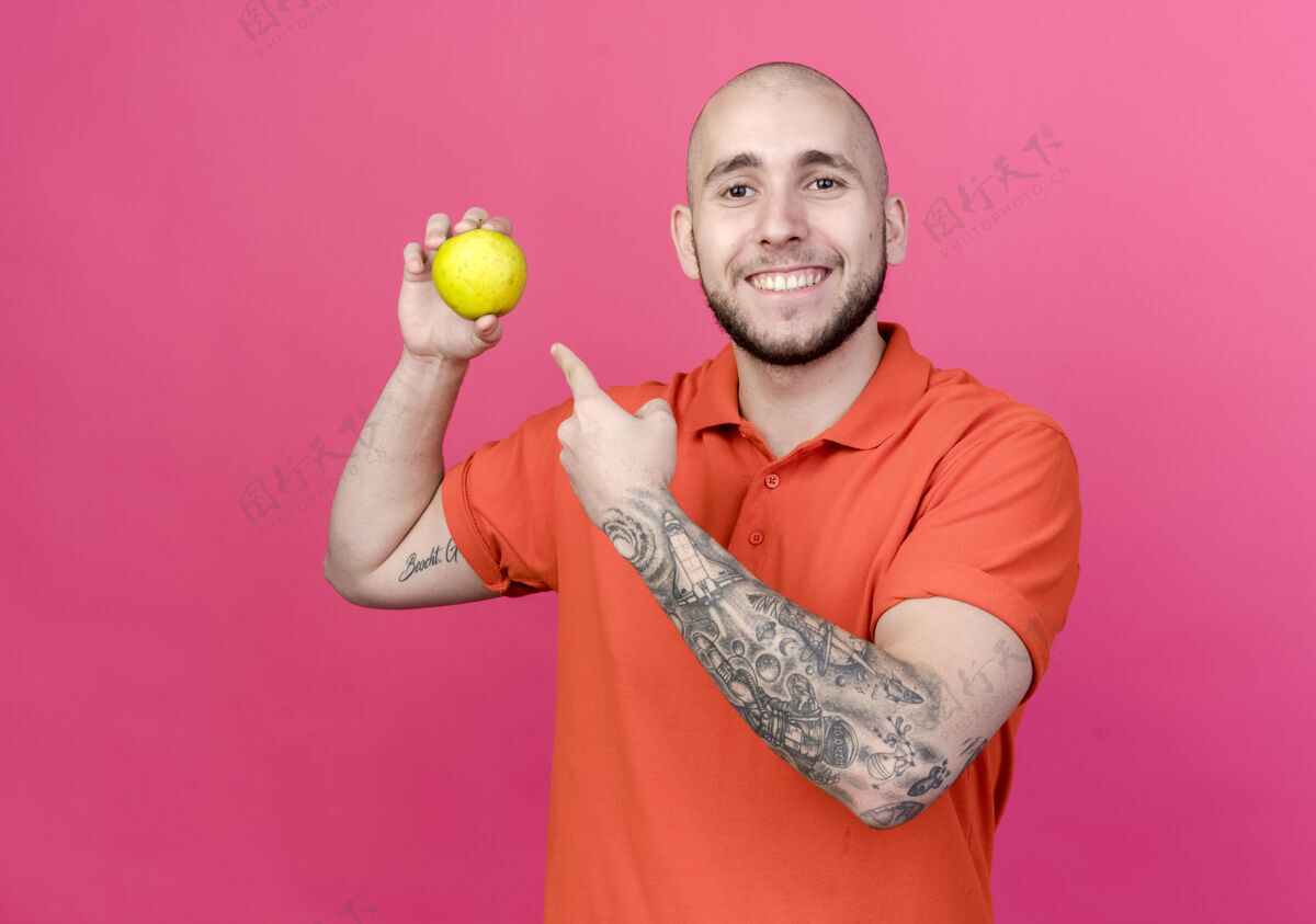 男人面带微笑的年轻运动型男子拿着苹果指着粉红色墙上的孤立点苹果微笑