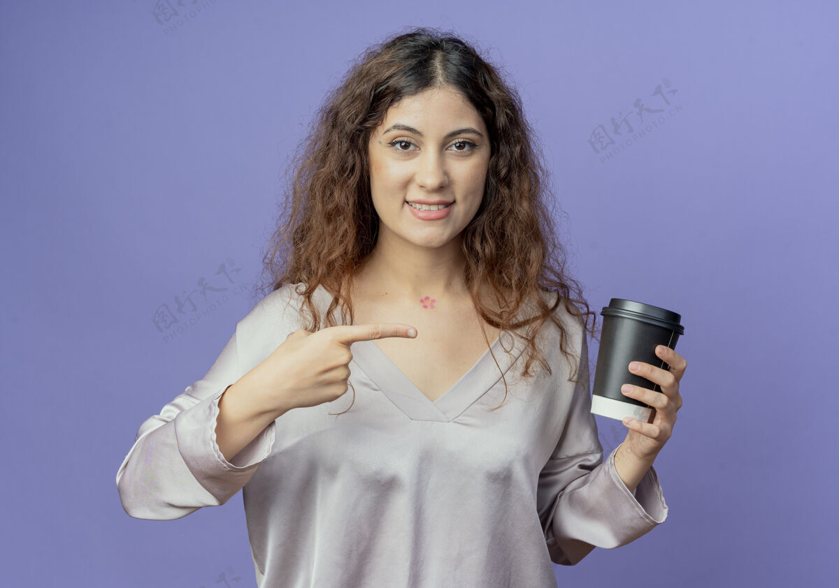 微笑微笑着的年轻漂亮的女孩拿着咖啡杯指着蓝色的墙壁女孩咖啡拿着