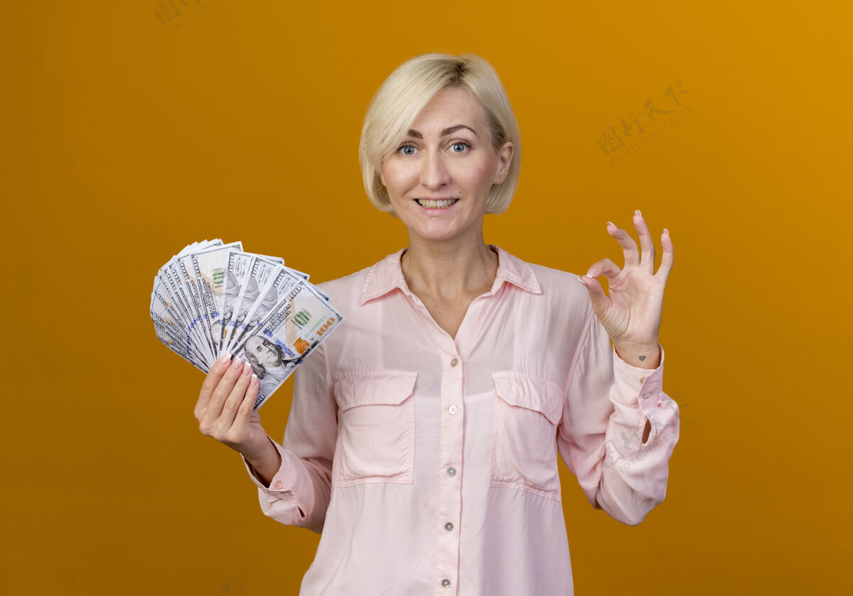 手势微笑着的年轻的金发斯拉夫女人拿着现金和显示奥基手势孤立在橙色墙上金发微笑斯拉夫人