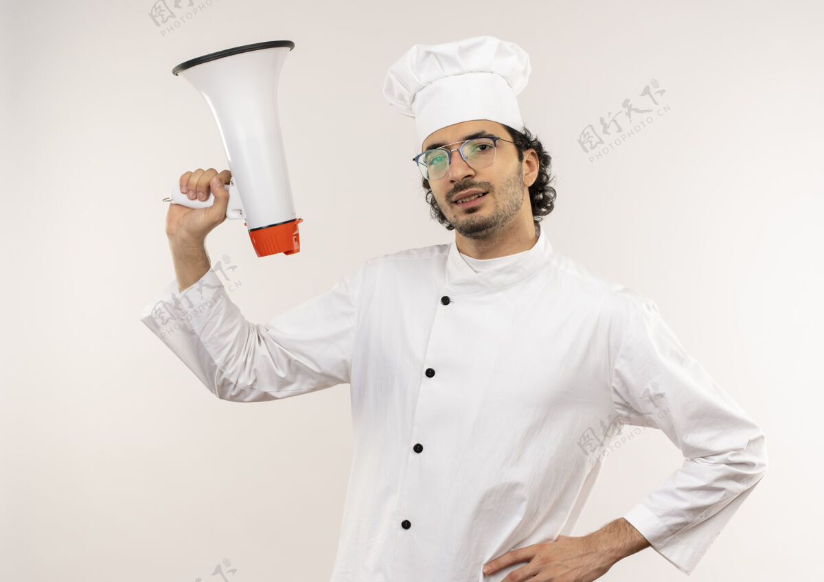 微笑面带微笑的年轻男厨师穿着厨师制服 戴着眼镜 举着扩音器 手放在臀部 隔离在白墙上提高臀部手