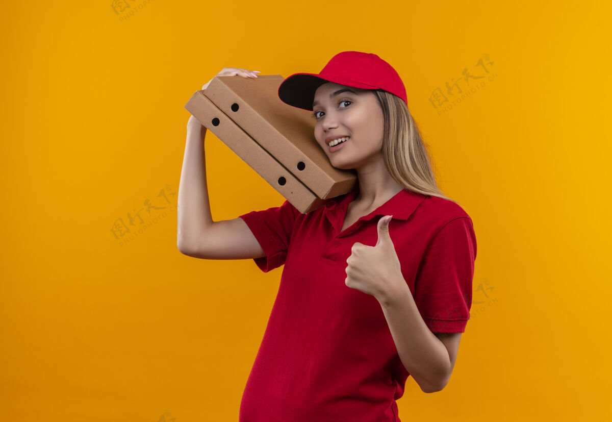 帽子微笑着的年轻送货女孩 穿着红色制服 戴着帽子 肩上扛着披萨盒 拇指孤立地竖立在橙色的墙上女孩拇指披萨