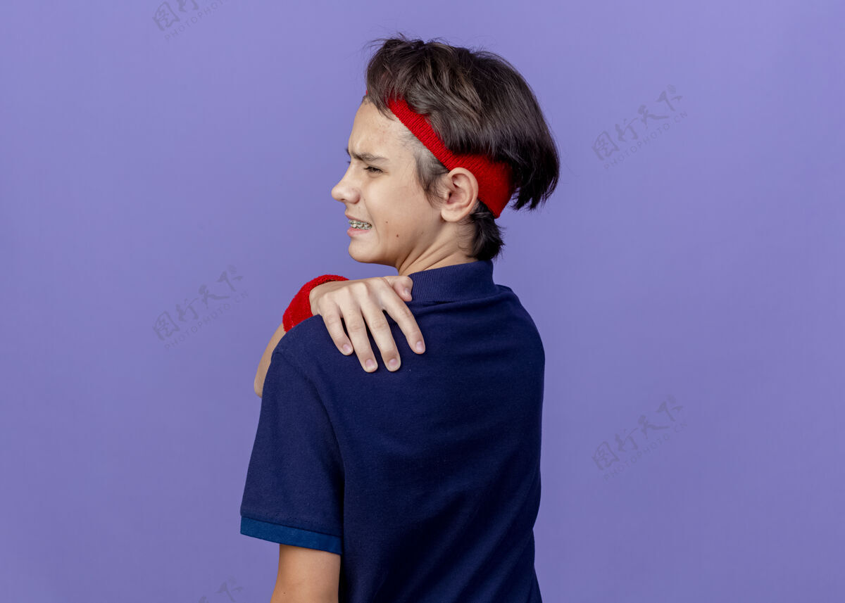 牙科疼痛的年轻英俊的运动男孩戴着头带和戴着牙套的腕带站在后面看侧面 手放在背上 隔离在紫色的墙上 留着复制空间运动背带腕带