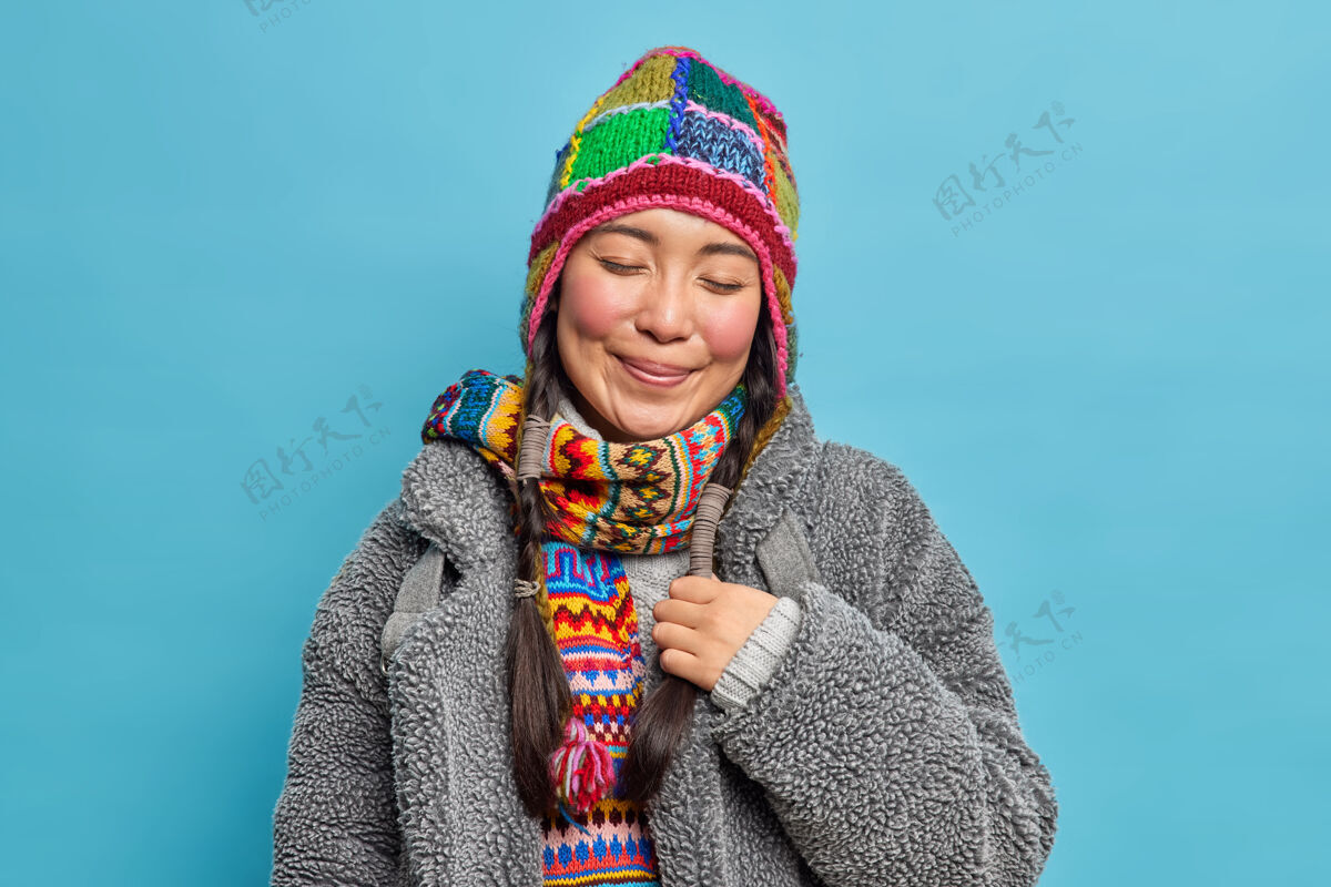 人类满意高兴的亚洲女人闭上眼睛 微笑愉快地戴着针织帽子和围巾寒冷的冬天温暖的外套有两个辫子回忆一些愉快的隔离在蓝色的墙上温暖人季节