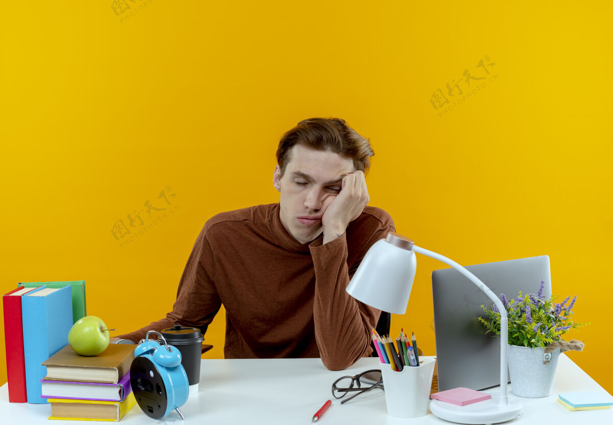 坐着一个疲惫的小男孩闭着眼睛坐在书桌旁 手里拿着工具 头戴着手腕 孤零零地躺在黄色的墙上学生累工具