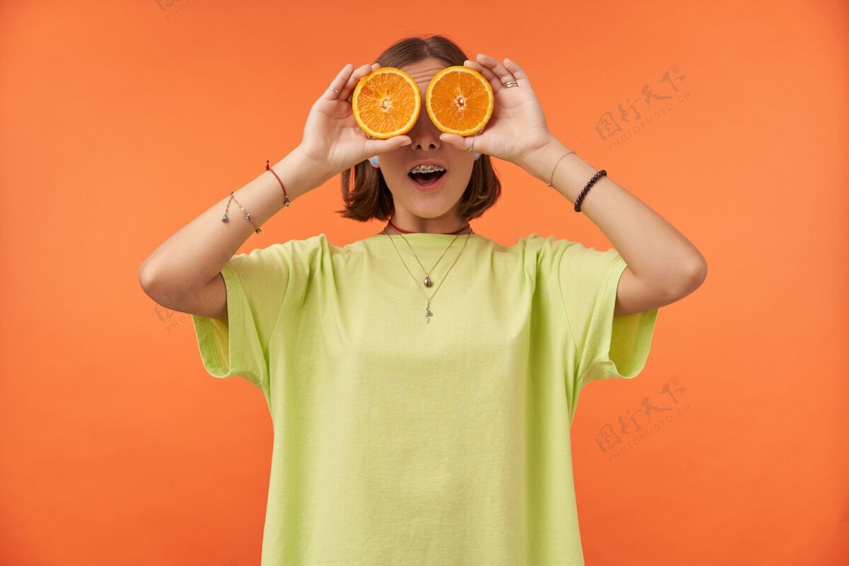 T恤女学生 一位棕发短发的年轻女士 眼睛上蒙着橘子神情惊讶站在橘色的墙上穿着绿色t恤 戴着牙套和手镯热带20多岁微笑