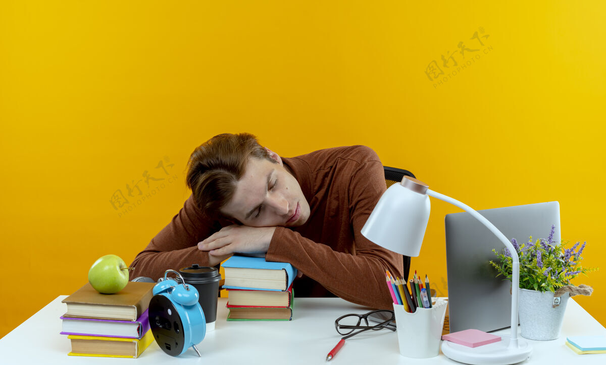 坐着年轻的学生男孩坐在书桌旁 手里拿着学习工具 手放在书上 孤零零地睡在黄色的墙上工具学生学校