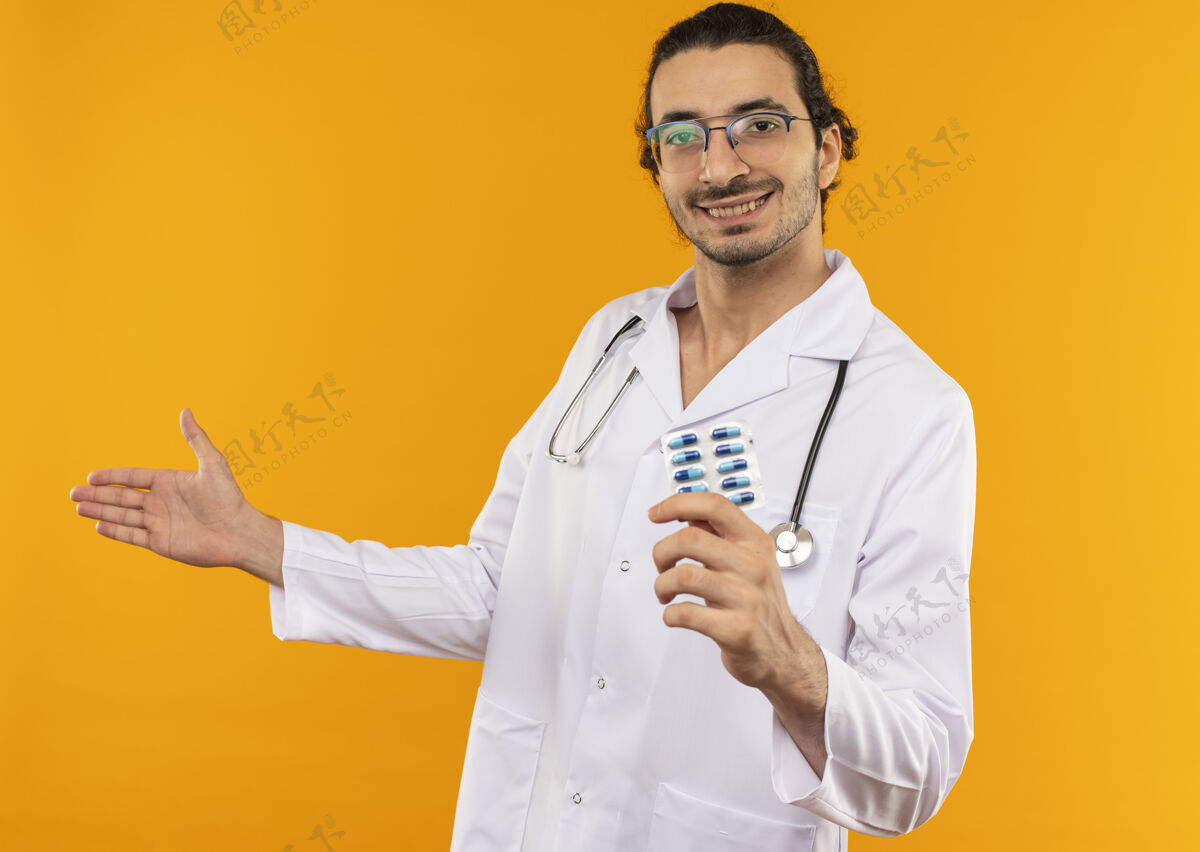 穿面带微笑的年轻医生 戴着医用眼镜 穿着医用长袍 手持听诊器 手持药片 用手指着隔离的黄色墙壁 并留有复印空间听诊器药丸医疗