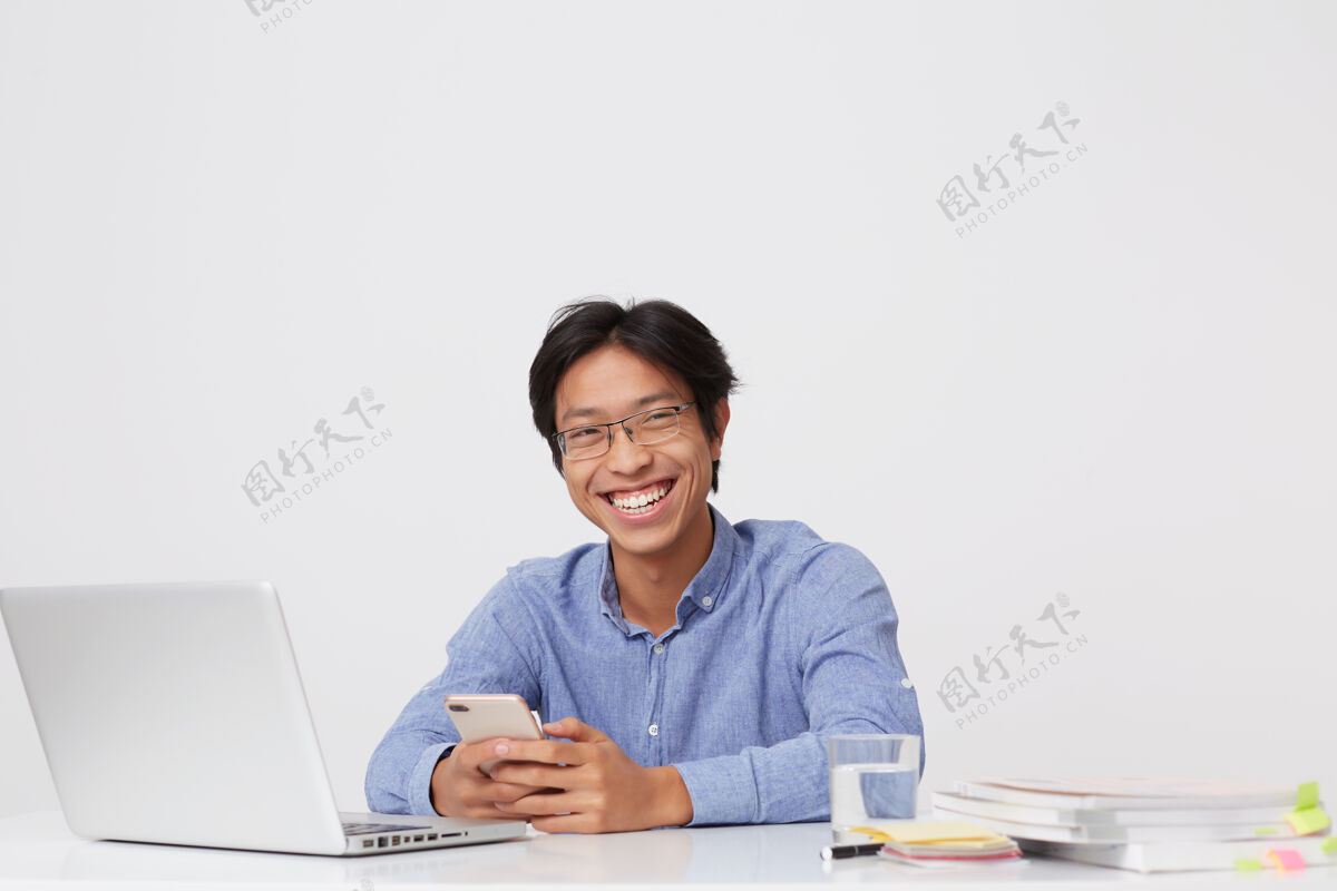 会议戴着眼镜的亚洲年轻商人 快乐地微笑着 一边用手机一边笑 一边用笔记本电脑在白墙上工作手机手机电脑