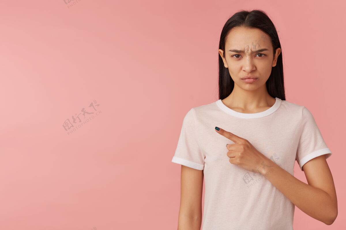 成人不高兴的 愤怒的女孩的肖像 黑色长发 穿着白色t恤 看着 皱着眉头手指指向左边的复制空间 隔离在粉彩粉色的墙上颜色漂亮情绪