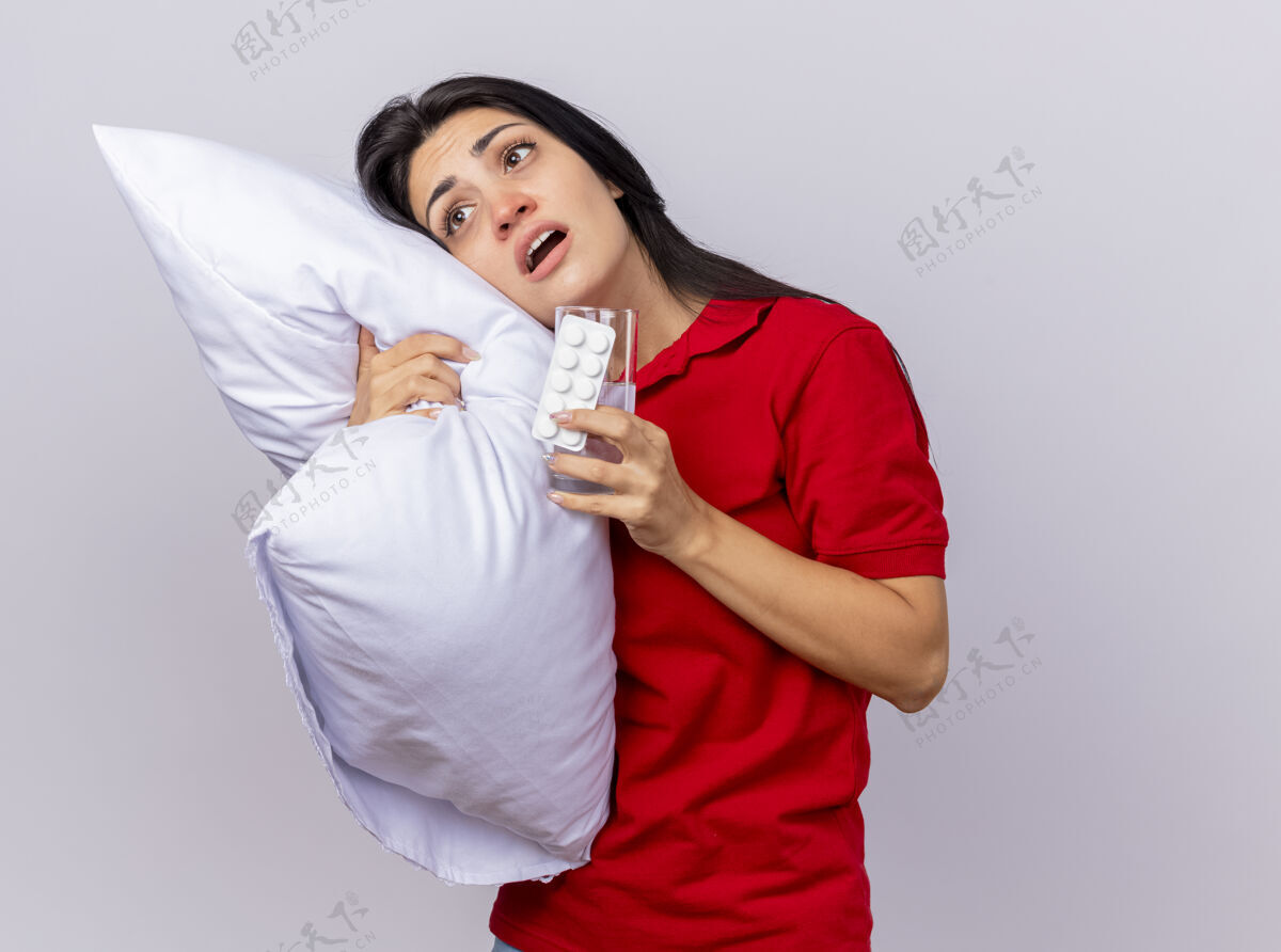 直令人印象深刻的年轻白种人生病的女孩抱着枕头把头放在枕头上 看着它与一包药片和一杯水在手上隔离在白色背景与复制空间直年轻包印象