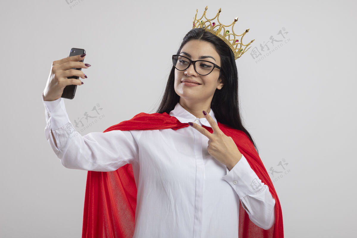皇冠高兴的年轻白种人超级英雄女孩戴着眼镜和王冠做和平标志采取自拍隔离在白色背景与复制空间年轻请拿自拍