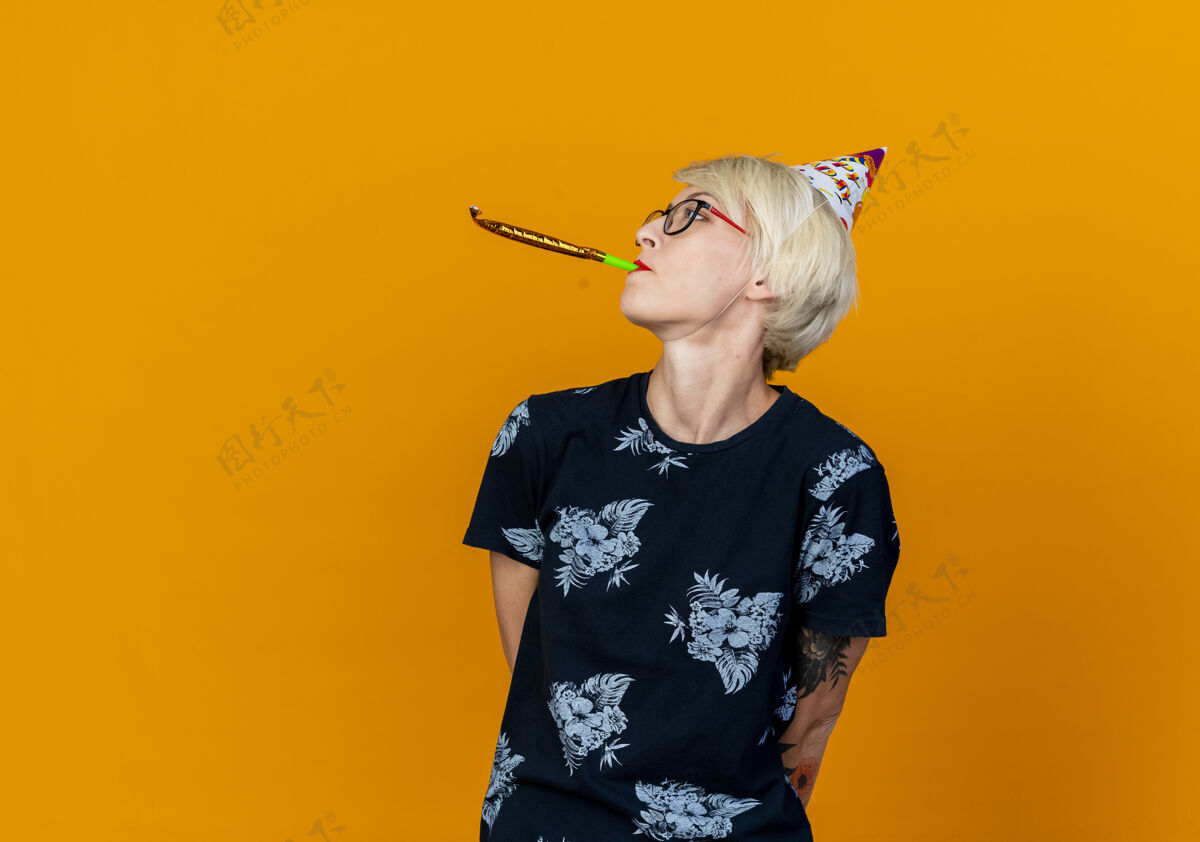 派对年轻的金发派对女郎戴着眼镜 戴着生日帽 双手放在背后 把头转向侧面吹派对风机 在橙色背景上隔离 留有复印空间眼镜侧板转