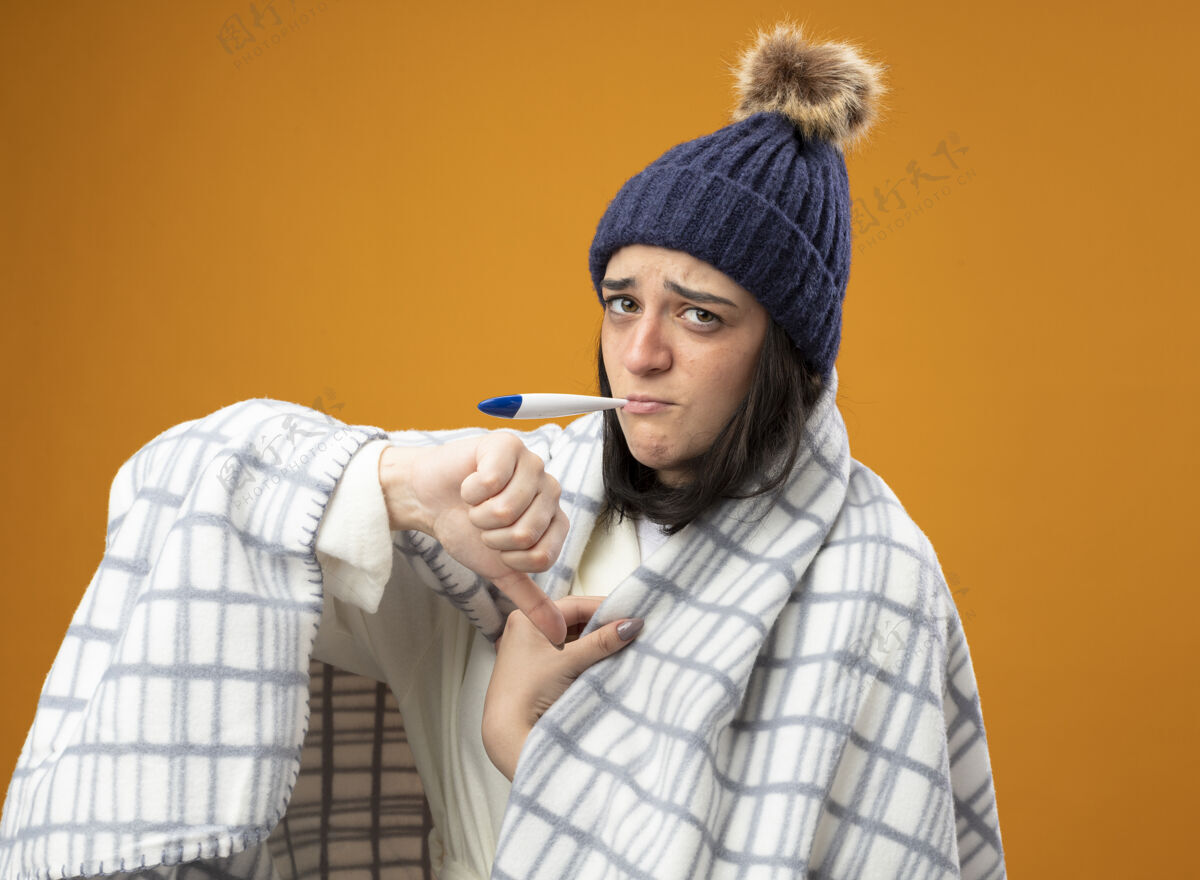 向下年轻的白种人生病的女孩穿着长袍 戴着格子呢的冬帽 看着摄像机 嘴里叼着温度计 拇指朝下 被隔离在橙色的背景上格子拇指长袍