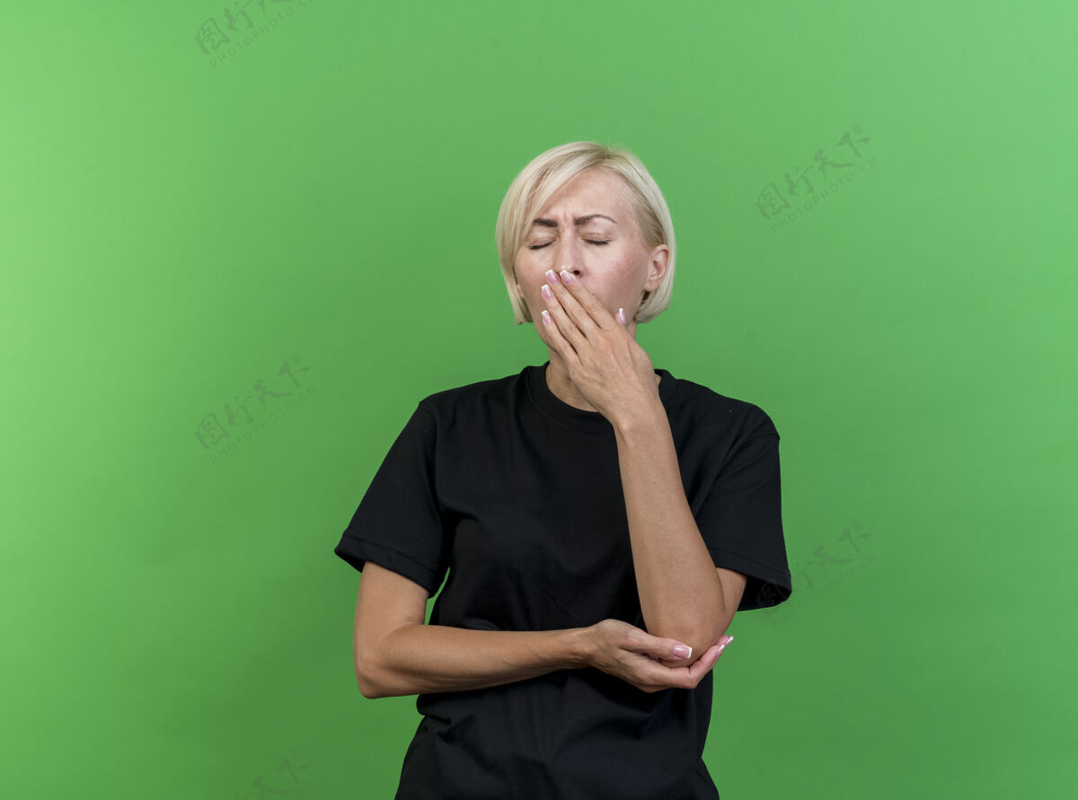 眼疲惫的中年金发斯拉夫女人把手放在手肘下打呵欠保持手放在嘴上闭着眼睛隔离在绿色背景与复制空间哈欠疲倦肘部