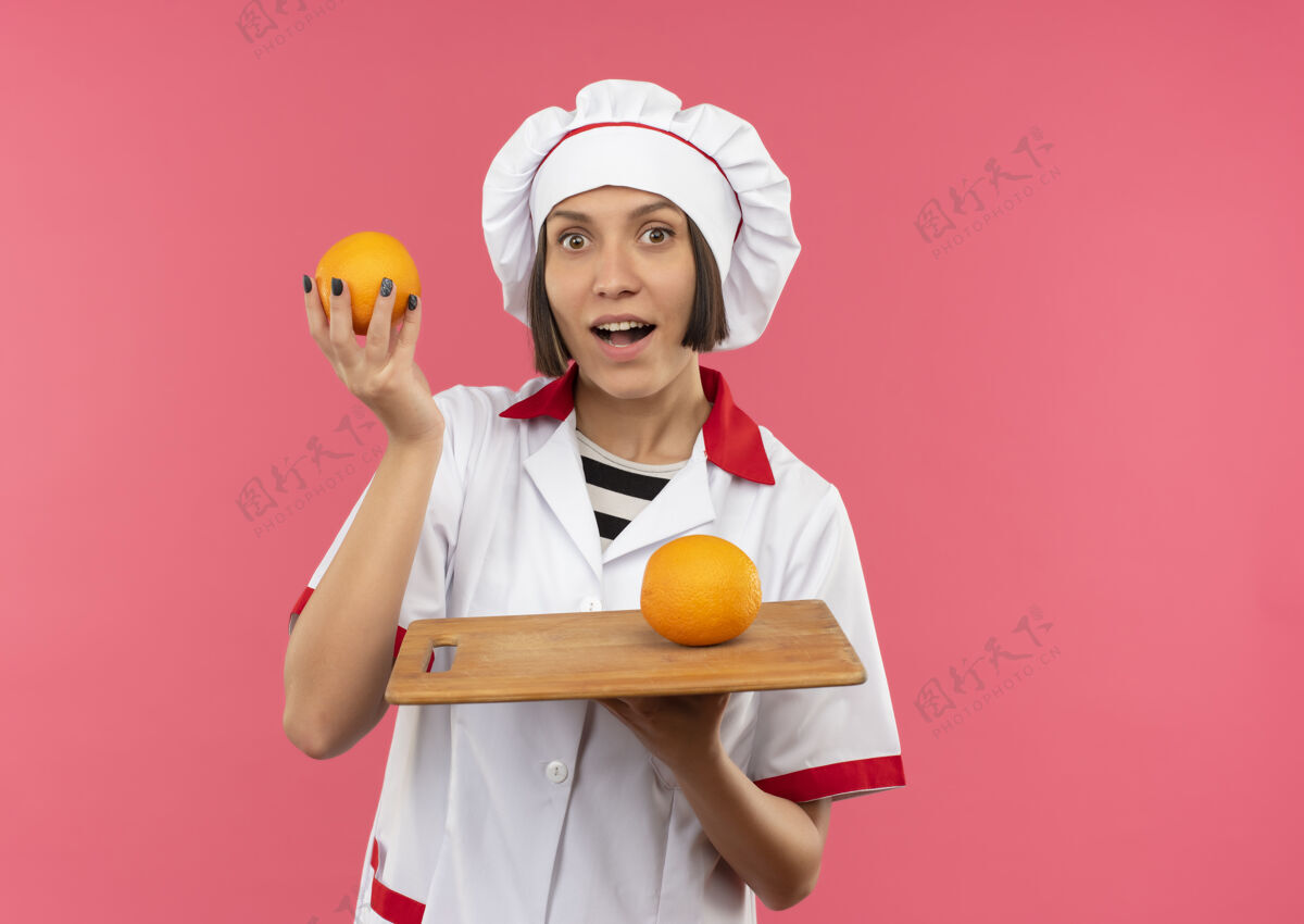 橙色令人印象深刻的年轻女厨师在厨师制服举行橙色和切菜板与橙色的它隔离在粉红色与复制空间烹饪空间女性