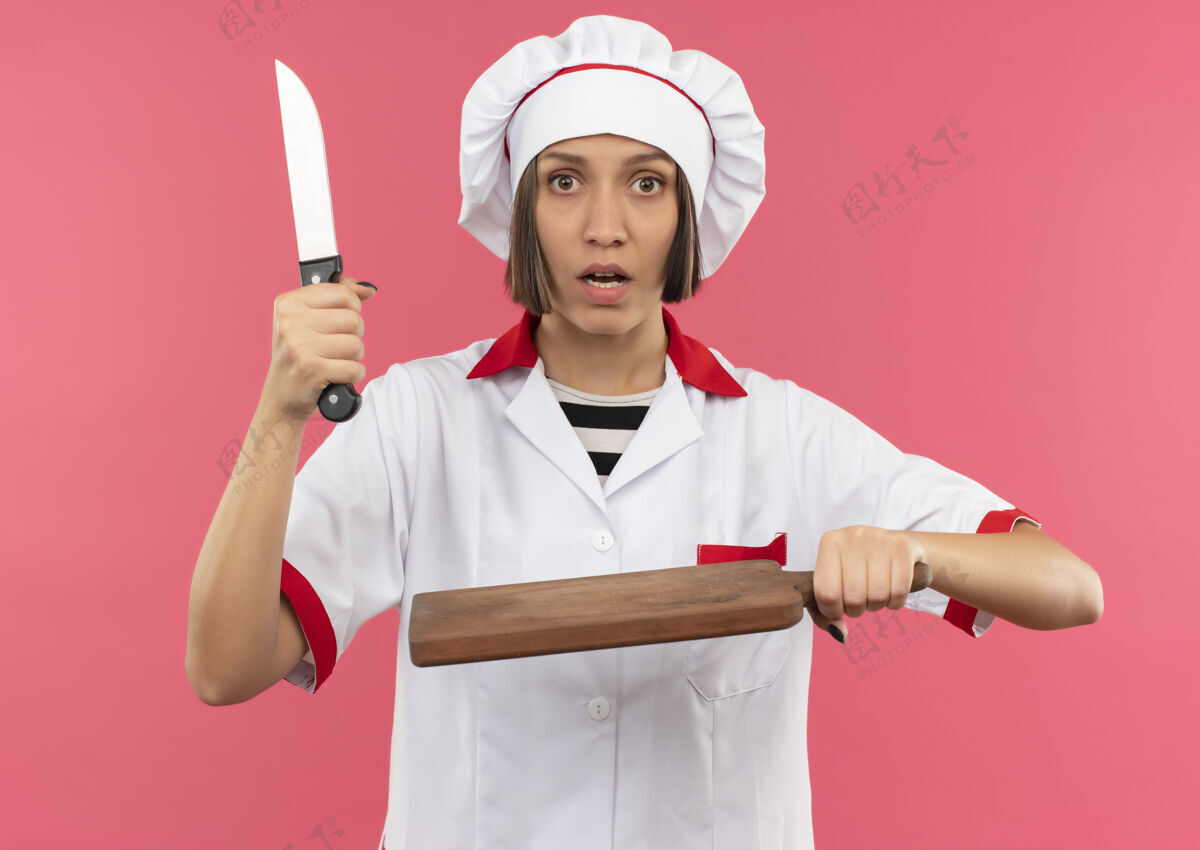 女印象深刻的年轻女厨师穿着厨师制服 手持刀和砧板上的粉红色隔离烹饪持有厨师
