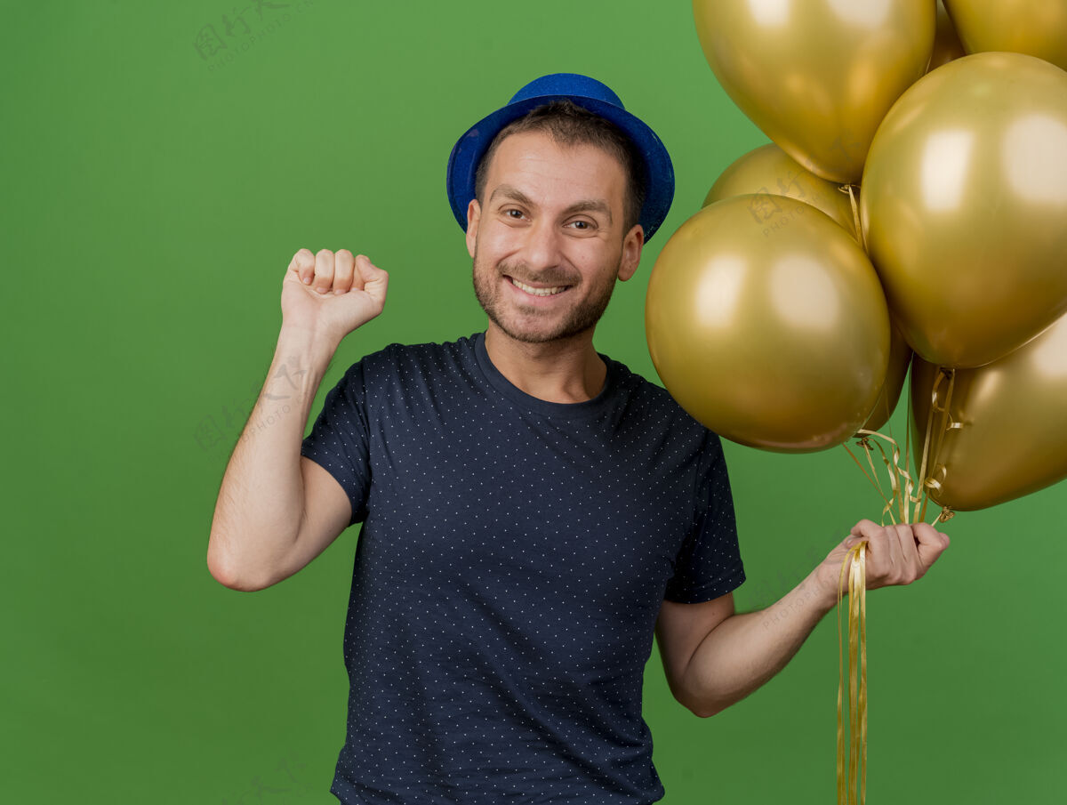 拳头面带微笑的白人帅哥戴着蓝色的派对帽 手里拿着氦气球 在绿色背景上举起拳头 留着复制空间派对持有气球