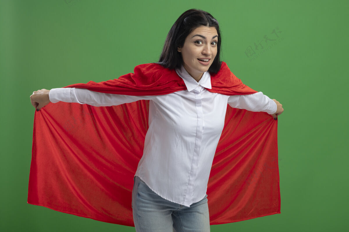 女孩顽皮的年轻白种人超级英雄女孩手持她的英雄斗篷 代表飞行 看着绿色背景上孤立的摄像机玩代表手持