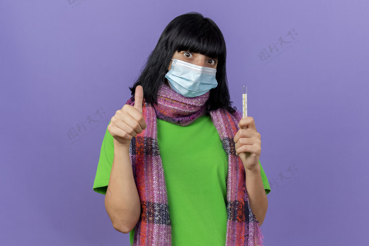 手握印象深刻的年轻病高加索女孩戴着面具和围巾拿着温度计看着相机显示拇指向上孤立的紫色背景与复制空间女孩体温计面具